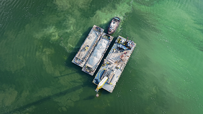 An aerial photo of a tug pushing a barge loaded with cobble in the center. An empty barge with a front-end loader sits to the left side. On the right side is a third barge with a tall crane and other construction equipment. The tug and barges are surrounded by calm, slightly muddy looking water.